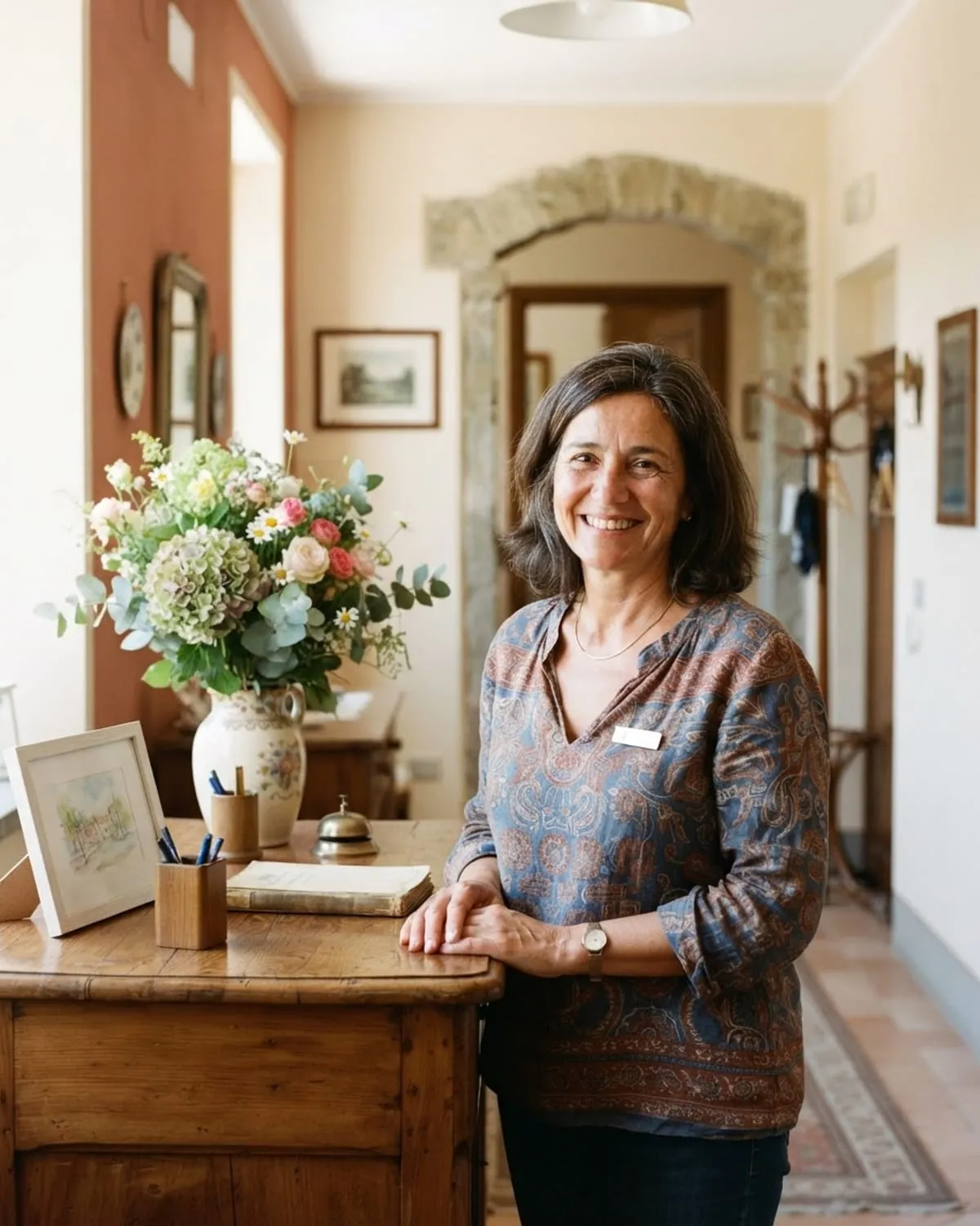 Titolare di B&B sorridente nell'ingresso luminoso, accanto a un vaso di fiori e al banco in legno